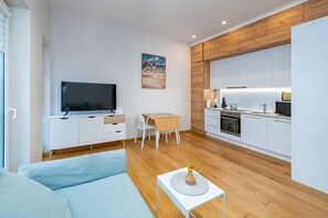 A modern living room with a flat-screen TV, a dining table, and a kitchen area with white cabinets and a wooden accent wall.