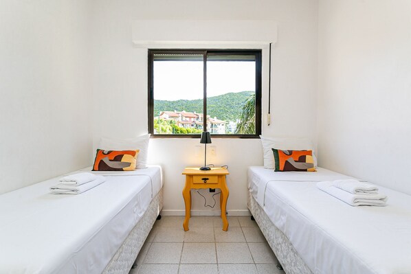 A room with two single beds, a small wooden nightstand with a lamp, and a window offering a view of buildings and greenery.