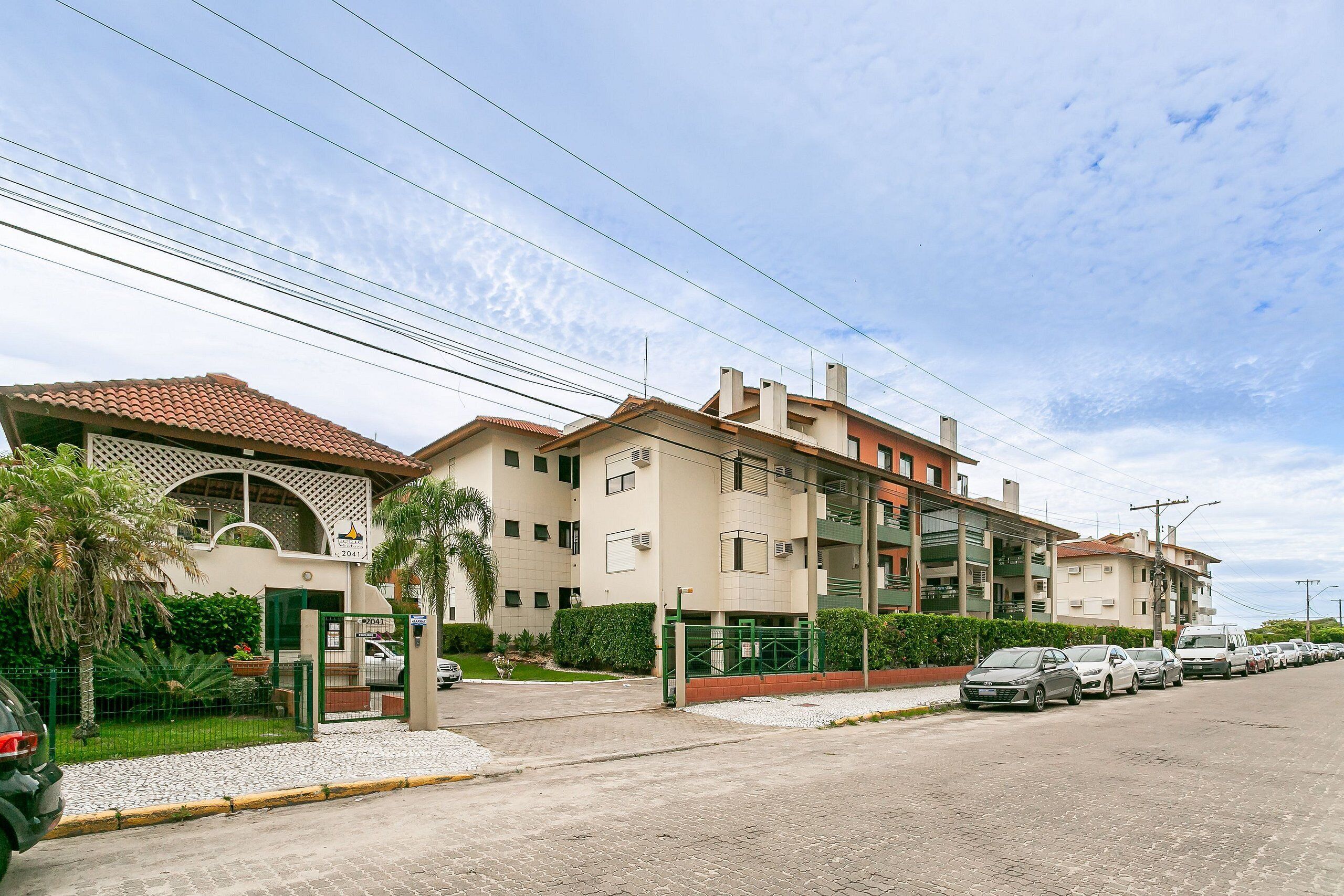 A multi-story residential building with a gated entrance, a parking area with several cars, and a clear sky.