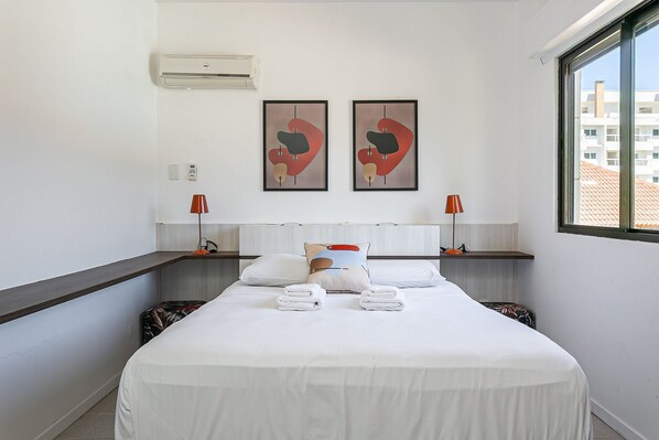 Habitación de hotel con una cama grande, dos aires acondicionados instalados en la pared, dos cuadros abstractos enmarcados, una mesita de noche con lámpara y una ventana con vistas a edificios.