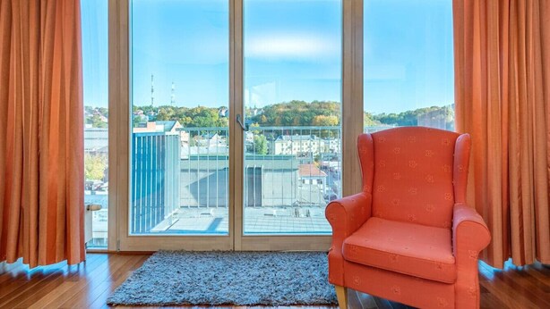 Una habitación con un gran ventanal corredero, un sillón rojo y vistas a edificios y árboles.