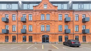 A multi-story brick building with large windows and balconies, a parking lot in front, and a car parked in the lot. - Shanti barracks ap by Polo apartments (Kaunas)