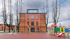 A playground with a slide and swings, a brick building with a large window, and bare trees. - Shanti barracks ap by Polo apartments (Kaunas)