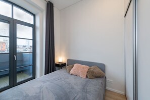 A bedroom with a bed, a nightstand, a lamp, and a view through a large window. - Shanti barracks ap by Polo apartments (Kaunas)