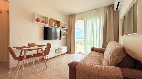 Una sala de estar moderna con televisor de pantalla plana, comedor con sillas de mimbre, sofá y ventana grande con vistas.