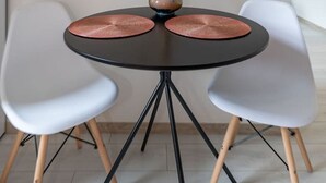 Uma mesa redonda com duas cadeiras brancas, um vaso com plantas secas e uma base preta.