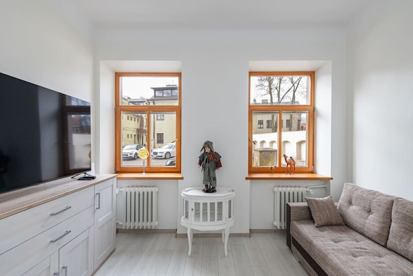 Un salon moderne avec un téléviseur, deux fenêtres à cadres en bois, un radiateur blanc, une table blanche et un canapé marron.
