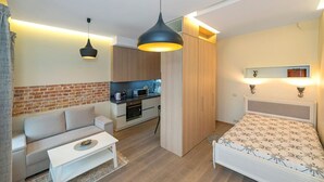 A compact room with a sofa, a small table, a bed, a kitchenette, and a hanging light fixture.