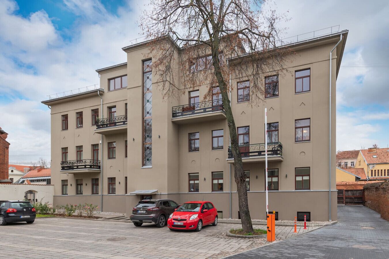 A multi-story building with balconies, a parking area with cars, and a tree in front.