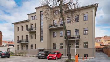 A multi-story building with balconies, a parking area with cars, and a tree in front.