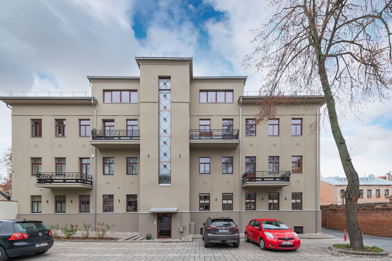 Un edificio de varios pisos con balcones, un elemento vertical central y automóviles estacionados.