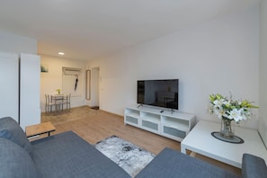 A modern living room with a flat-screen TV, grey sofas, a coffee table, a dining area with chairs, and a vase with flowers.