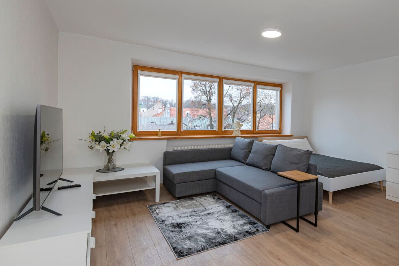 Uma sala de estar moderna com sofá seccional, TV de tela plana, mesinha branca e uma grande janela com vista para edifícios.