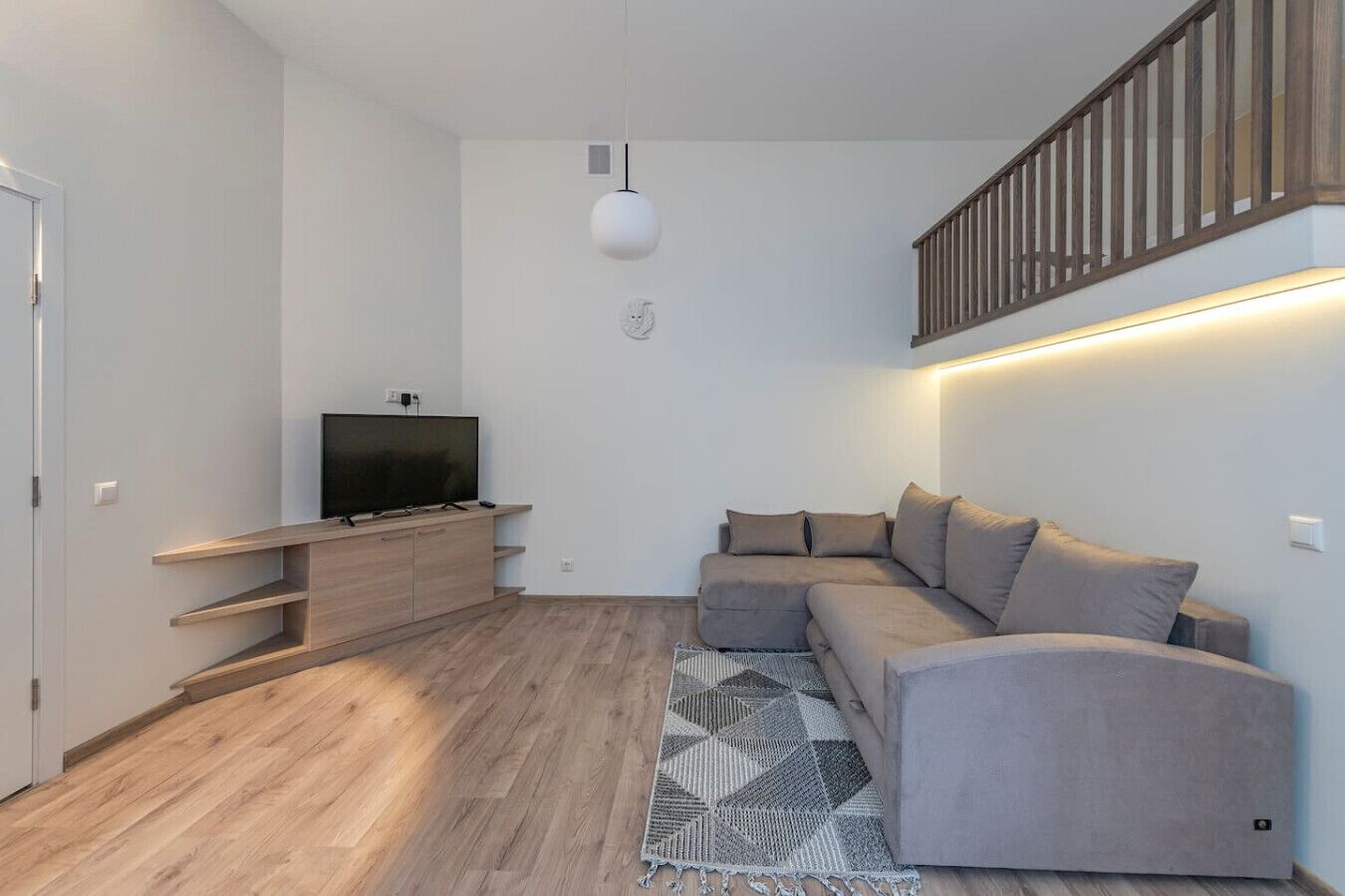 Ett modernt vardagsrum med en platt-TV placerad på en träkonsol, en grå soffa, en mönstrad matta och en trappa med träräcken.