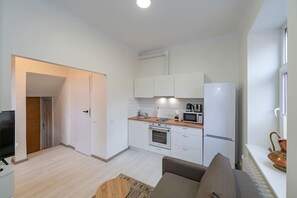 Eine kompakte Küche mit weißen Schränken, einer Mikrowelle, einem Backofen und einem Kühlschrank. Im Wohnbereich befinden sich ein Holzboden, eine Couch und ein Fernseher.