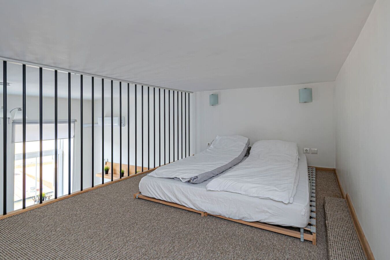 A bedroom with a bed, a view through a window with vertical blinds, and a small plant on the windowsill.