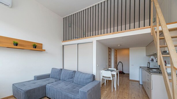 Een modern appartement met een grijze modulaire bank, een kleine eettafel, een kitchenette en een houten trap.