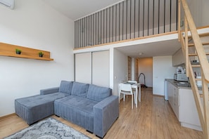 Un apartamento moderno con un sofá seccional gris, una pequeña mesa de comedor, una kitchenette y una escalera de madera.