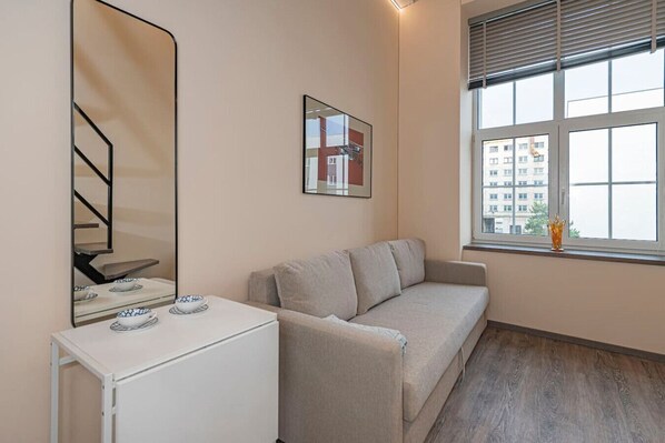 Une pièce minimaliste comprenant un canapé, une petite table avec un bol, un miroir et une fenêtre donnant sur des immeubles.