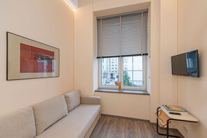 Ein Zimmer mit einer Couch, einem Fenster mit Jalousien, einem Flachbildfernseher und einem kleinen Tisch mit Büchern und einer Fernbedienung.
