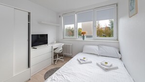 Ein Schlafzimmer mit Bett, Schreibtisch mit Fernseher, Heizkörper und Fenster mit Blick auf Bäume und Gebäude.
