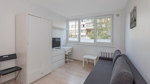 Ein kleines Zimmer mit Bett, Schreibtisch, Stuhl, Kleiderschrank und einem Fenster mit Blick auf Gebäude.