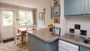 A kitchen with a built-in dishwasher, a dining area with a table and chairs, and a window with curtains. - Sunny Side (Saltburn-by-the-Sea)