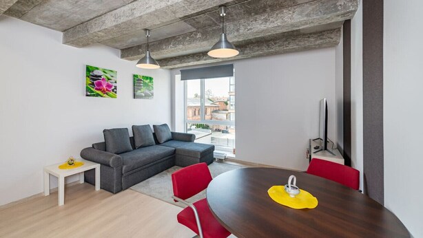A modern living space with a grey sectional sofa, a wooden dining table with red chairs, a small white side table, and a large window offering a view of buildings outside. - Ap Accent by Polo apartments (Kaunas)