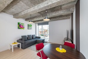 A modern living space with a grey sectional sofa, a wooden dining table with red chairs, a small white side table, and a large window offering a view of buildings outside.