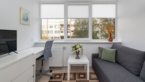 Un salon moderne avec un bureau, un canapé gris, une table basse blanche et une fenêtre équipée de stores.