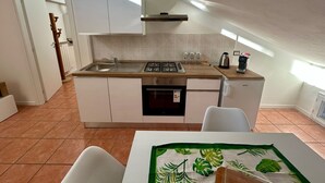 A compact kitchen with white cabinets, a sink, a stove, and a dishwasher. There is a window providing natural light, and a chair is visible in the foreground. - Stefano's cozy apartment (Erba)