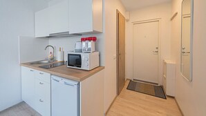 Cocina compacta con microondas, tostadora y lavabo, un refrigerador pequeño, y una puerta que da a un baño con espejo y ducha.