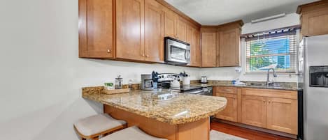 Una cocina con armarios de madera, encimera de granito, refrigerador de acero inoxidable y una ventana con persianas.