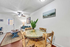Un coin repas avec une table ronde en verre, des chaises en bois, un ventilateur de plafond, une plante en pot et un tableau accroché au mur.