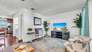 A living room with a view of the beach, a wooden coffee table, a patterned armchair, and a side table with a lamp. - Sandpiper Philosophy (Panama City Beach)