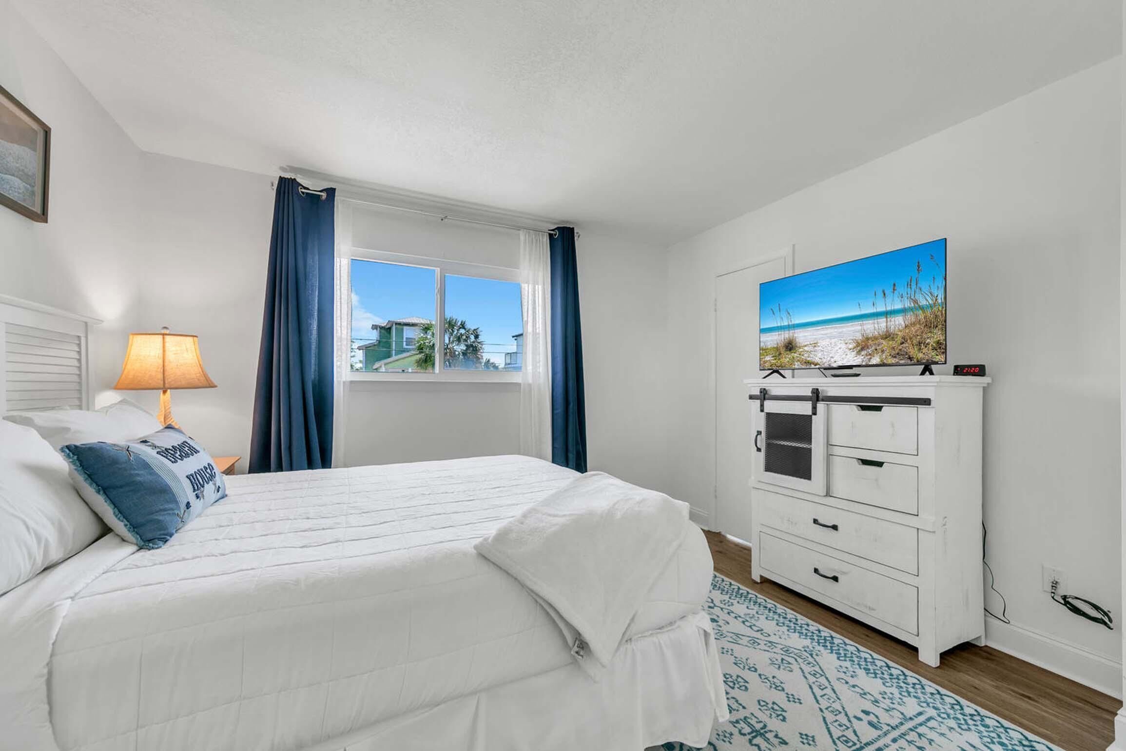A bedroom with a bed, a nightstand, a lamp, a dresser, a window with curtains, and a painting of a beach scene.