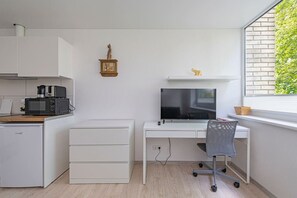 A compact kitchen with a microwave, toaster, and coffee maker, a white desk with a TV, and a window with a brick wall and greenery.
