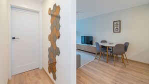Ein modernes Zimmer mit einer weißen Tür, einem an der Wand befestigten abstrakten Kunstwerk, einem Essbereich mit Tisch und Stühlen sowie einem Fernseher auf einer niedrigen Ablagefläche.