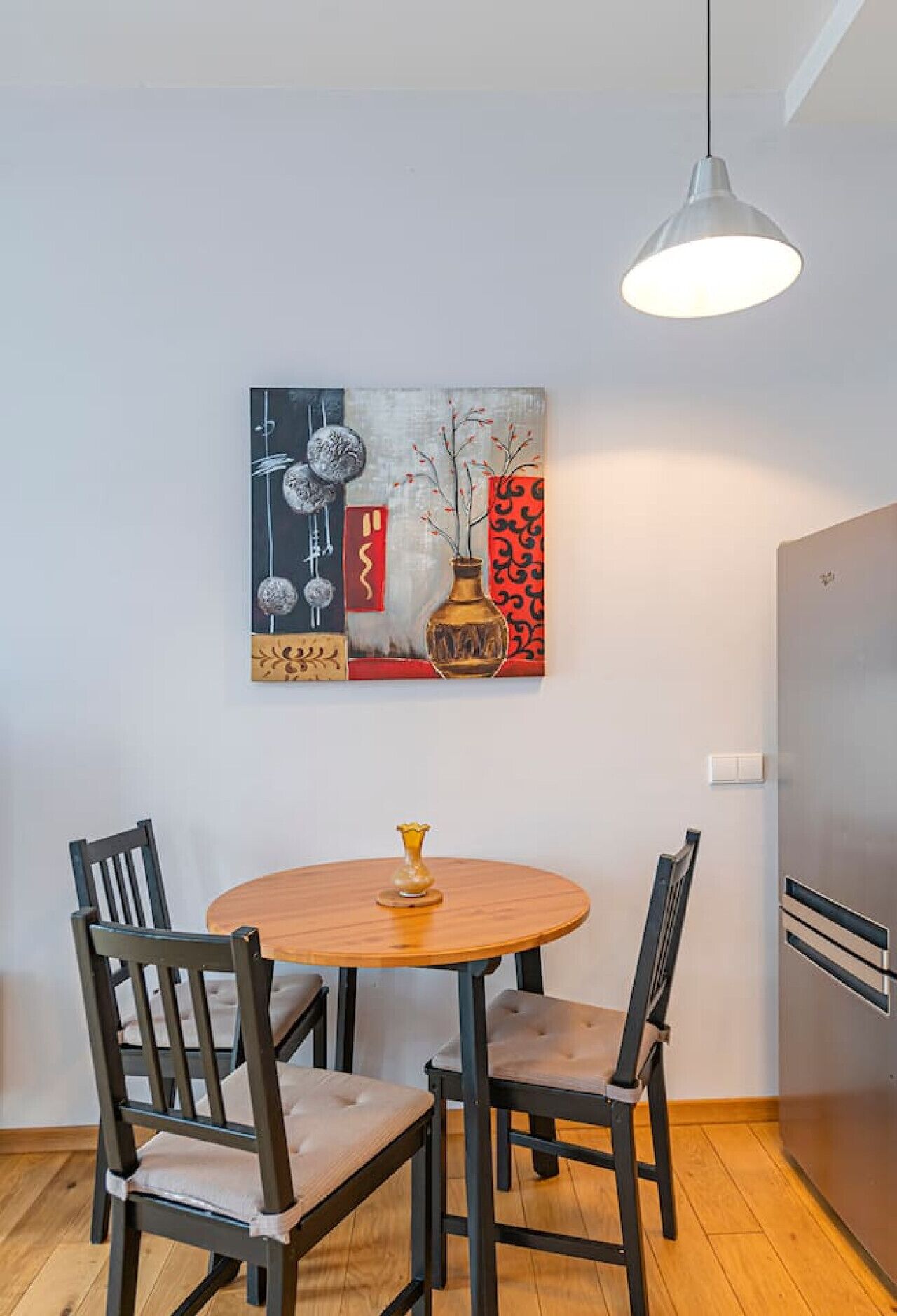 Un coin repas comprenant une table ronde en bois, quatre chaises, une suspension et un tableau accroché au mur.