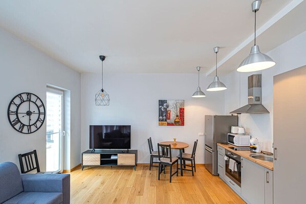 Una sala de estar moderna con televisor de pantalla plana, una zona de comedor con una mesa redonda y sillas, una cocina con refrigerador y microondas, y un reloj de pared grande.