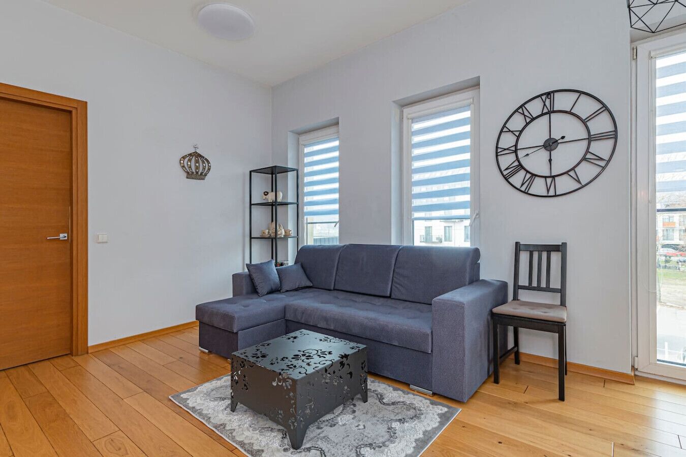 A living room with a grey sofa, a small coffee table, a bookshelf, a chair, and a wall clock.