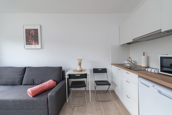 A compact living space with a grey sofa, a small dining table, two chairs, a kitchen area with a microwave, and a single framed picture on the wall.