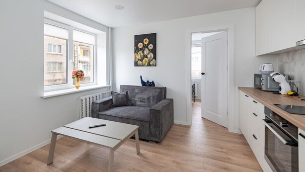A modern apartment with a grey sofa, a small table, a kitchen with a coffee machine, and a window with a view of buildings.