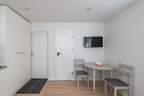 A small, clean room with a white door, a television mounted on the wall, a round table with two chairs, and a small rug on the floor. - Ap Dandelion by Polo apartments (Kaunas)