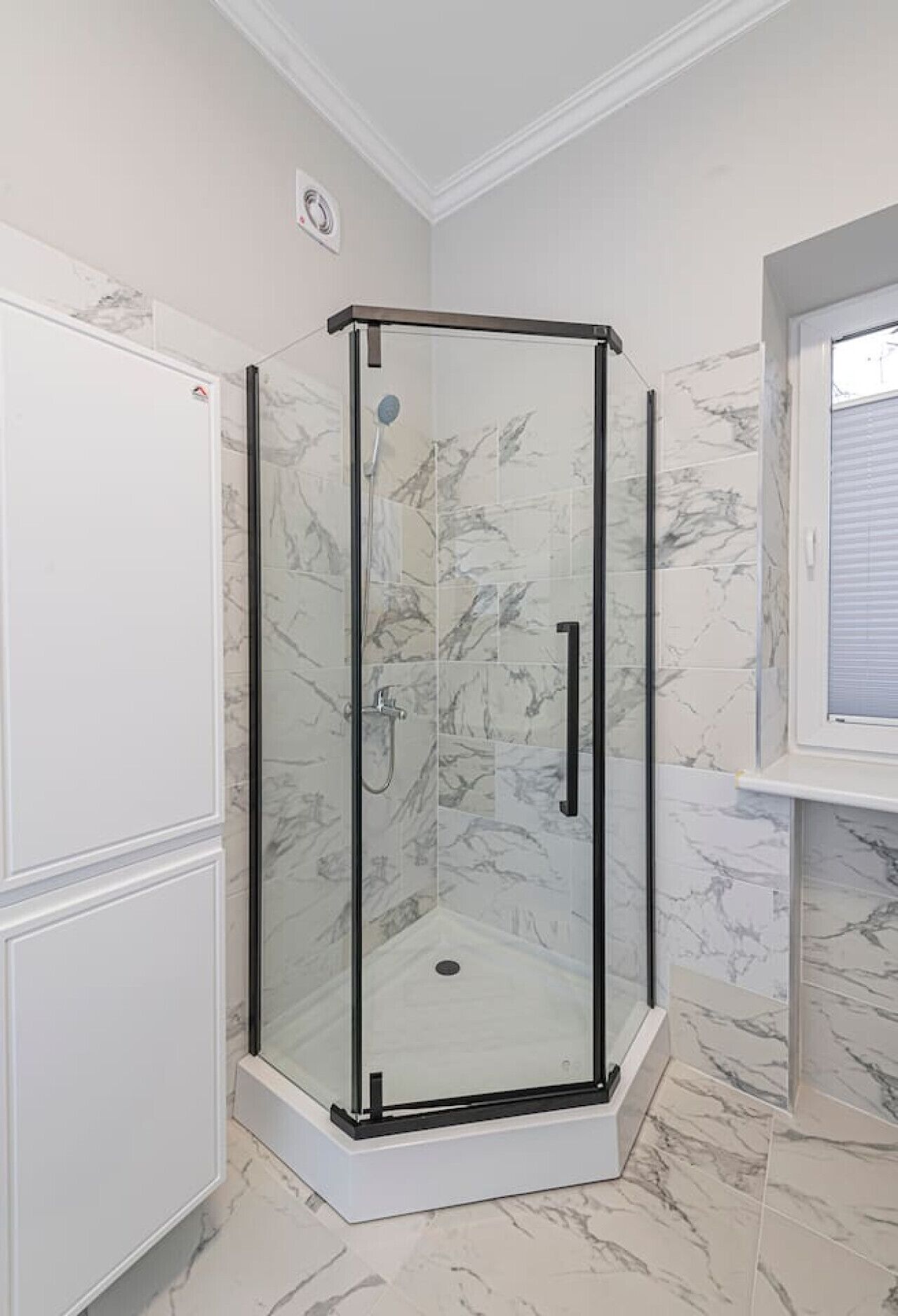 A corner shower enclosure with glass doors, a white tiled floor, and marble wall tiles.