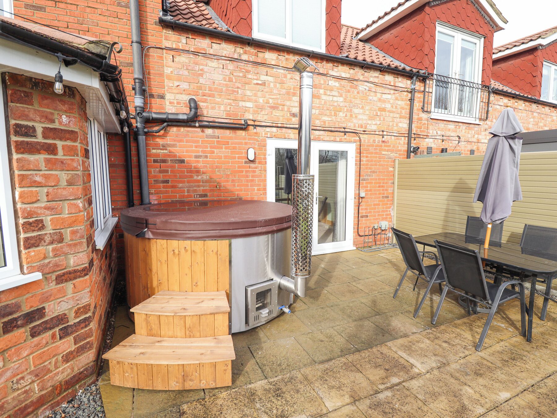 A hot tub with wooden steps, a table with chairs, and a brick wall.