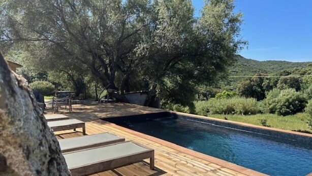 Una terraza de madera con piscina, rodeada de árboles y un paisaje montañoso.