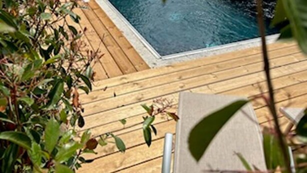 A wooden deck with a swimming pool, surrounded by greenery and a view of hills in the background.