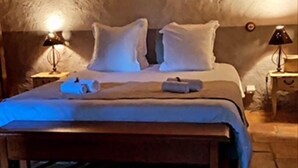 Un dormitorio acogedor con techo de madera, pared de piedra y cama con ropa blanca. Hay dos mesitas de noche con lámparas.