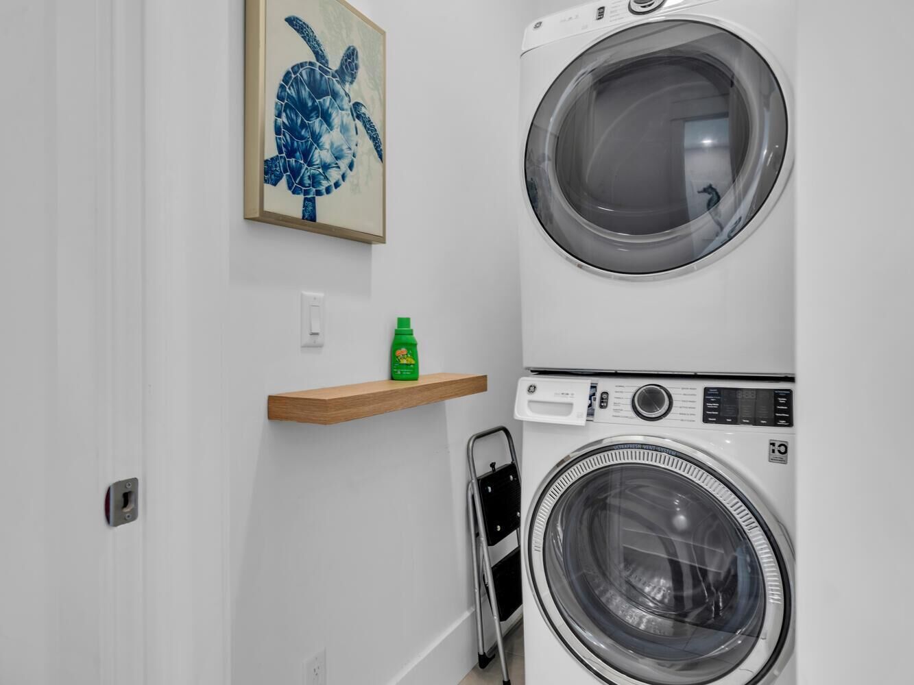 A laundry area with stacked washers and dryers, a wooden shelf with a bottle, and a framed artwork on the wall.